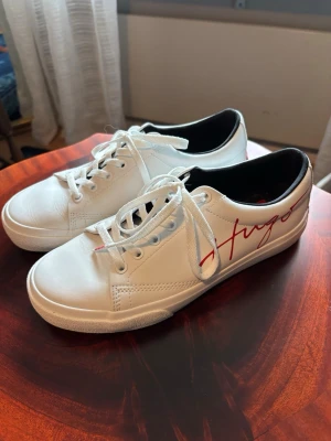 Vita sneakers från Hugo med röd logga - Snygga vita sneakers från Hugo med röd logga på sidan och vita snören. Skorna har en klassisk låg profil och svart innerfoder. Tillverkade i slätt läderliknande material och har en stilren look som passar till det mesta.