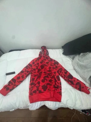 Röd camo hoodie med tryck - Säljer en riktigt fet röd hoodie med svart camomönster och coolt tryck på ärmen. Hoodien har dragkedja, fickor och en stor huva. Perfekt för dig som vill sticka ut med streetwear vibes. Materialet känns mjukt och skönt, passar till chill eller när du vill ha en statement look.