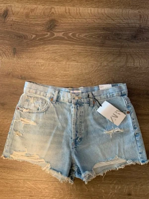 Ljusa Low denimshorts från Zara - Säljer ett par ljusblå denimshorts från Zara med råa kanter och slitna detaljer. Klassisk femficksmodell med hög midja och knappgylf. Perfekta för sommaren och ger en avslappnad vibe till din outfit.