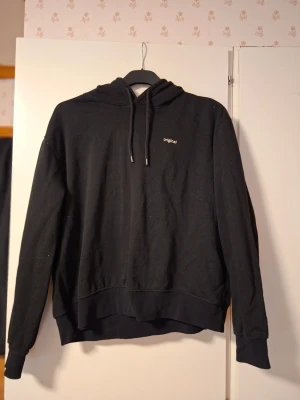 Svart hoodie med tryck 'original' - En svart hoodie med huva och dragsnören. Liten vit text 'original' på bröstet. Klassisk passform med ribbade muddar vid ärmslut och nederkant.
