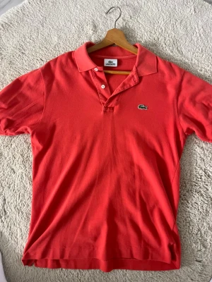 Orange pikétröja från Lacoste - Klassisk orange pikétröja från Lacoste med korta ärmar och den ikoniska gröna krokodilloggan på bröstet. Tröjan har krage och knappar framtill. Perfekt för en avslappnad och stilren look.