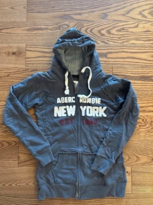 Marinblå hoodie Abercrombie and fitch  - Snygg marinblå hoodie från Abercrombie med dragkedja och broderad text 'NEW YORK' på bröstet. Har huva med vita snören och kängurufickor framtill. Detaljer i vitt och rött ger en cool kontrast. Perfekt för en avslappnad streetstyle-look.