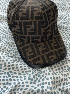 Fendi FF-logga keps brun/svart - Säljer en snygg keps från Fendi med klassiskt FF-monogram i brunt och svart. Kepsen har böjd skärm med svart kant och justerbart band bak i läder med Fendi-logga. Perfekt för dig som gillar exklusiva accessoarer och streetstyle.