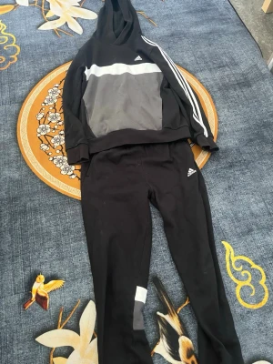 Svart Adidas loungewear set - Snyggt svart loungewear set från Adidas med hoodie och byxor. Tröjan har vit och grå panel framtill samt klassiska vita ränder på ena ärmen. Byxorna har vita och grå detaljer vid benen samt Adidas-logga. Perfekt för chill dagar hemma.