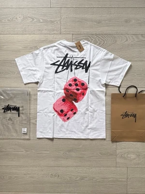Stussy / Stüssy t-shirt - Helt ny t-shirt från Stussy med svart logga på bröstet och  tryck av två rosa tärningar samt logga på ryggen. Klassisk passform och korta ärmar. Perfekt för sommaren och dig som gillar streetwear. Papperspåse och plastpåse ingår. Priset går att diskutera. Kom gärna med frågor!😁