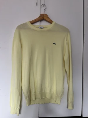 Ljusgul tröja från Lacoste - Snygg ljusgul tröja från Lacoste med klassisk rund halsringning och den ikoniska gröna krokodilloggan broderad på bröstet. Tillverkad i mjukt, tunt material som känns skönt mot huden. Perfekt för dig som gillar stilrena och tidlösa plagg.