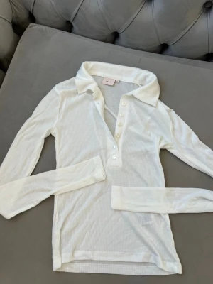 Vit ribbad långärmad topp Nelly - Säljer en vit ribbad långärmad topp från Nelly med klassisk krage och knappar framtill. Toppen har en slim passform och är tillverkad i mjuk bomullsmix som känns skön mot huden. Perfekt att styla med jeans eller kjol för en clean look. Materialet är lite genomskinligt för en trendig look. Toppen är helt ny och oanvänd!
