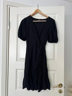 Somrig omlottklänning Gina Tricot - Svart omlottklänning från Gina Tricot med puffärmar och volangkant nertill. Klänningen har knyt i midjan och v-ringad hals. Perfekt för dig som gillar en enkel men trendig look.