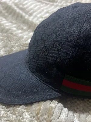 Svart Gucci keps med GG-mönster - Säljer en svart keps från Gucci med det klassiska GG-monogrammet över hela kepsen. På sidan finns en grön och röd rand som ger den en ikonisk look. Kepsen har böjd skärm och är tillverkad i ett slitstarkt tygmaterial.
