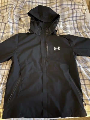 Svart vindjacka från Under Armour - Snygg svart vindjacka från Under Armour med huva och dragkedja. Jackan har två sidofickor med dragkedja och logga på bröstet. Tillverkad i vindavvisande material, perfekt för regniga och vindiga dagar. Sportig design med justerbar huva och detaljer i vitt.