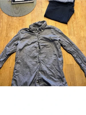 Grå skjorta med knappar från Gant - Klassisk grå skjorta från Gant med button-down krage och långa ärmar. Skjortan har knäppning framtill och en liten broderad logga på bröstet. Perfekt till jeans eller chinos för en stilren look.