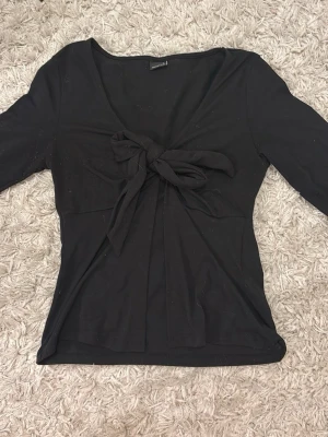 Svart topp med knyt från Gina Tricot - Svart långärmad topp från Gina Tricot med djup v-ringning och snygg knytning framtill. Toppens material är mjukt och stretchigt, vilket gör den bekväm och enkel att styla till olika looks.