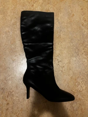 Svarta höga stövlar med klack - Säljer ett par svarta höga stövlar smal klack. Stövlarna är i glansigt konstläder och har en slouchy passform på skaftet. Perfekta för dig som vill ha en edgy och trendig look. Pris kan alltid diskuteras💖
