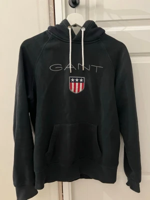 Svart hoodie från GANT - Svart hoodie från GANT med broderad logga och sköld på bröstet. Klassisk känguruficka framtill och vita dragsnören i huvan. Har lite skav ringen vid kragen (se bild) 
