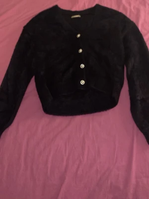 Kofta ifrån Only köpt i Åhléns - Mysig svart kofta i fluffigt material storlek 146-152 med långa ärmar och v-ringning om man har på alla knappar. Man kan ta av och på knapparna. Fyra dekorativa pärlknappar med glittrande kant. Perfekt för dig som vill ha en trendig och elegant look.