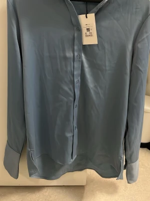 Blå satinliknande skjorta med knappar - Snygg blå skjorta i satinliknande material med klassisk krage och knappar framtill. Långärmad modell med breda manschetter och knappar vid ärmslut. Är lite stor eller översized så passar nog bäst M, kanske även större, ärmarna är också långa men går att vika in, blir dock rätt så tajt. Modellen är längre bakifrån och har två små slitsar på sidorna