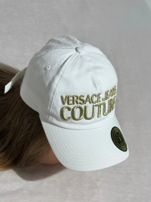 Vit keps Versace Jeans Couture - Helt ny , oanvänd och snygg vit keps från Versace Jeans Couture med broderad logga i guld framtill. Klassisk böjd skärm och justerbar rem bak. Tillverkad i bomull för en bekväm och luftig känsla. Perfekt för dig som vill ha en stilren accessoar med lyxig touch. Utan prislapp
