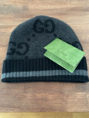 Gucci mörkgrå mössa med GG-logga - Snygg mörkgrå mössa från Gucci med svart GG-logga och ribbad kant med grå rand. Tillverkad i mjukt material som känns lyxigt och passar perfekt till höst och vinter. En stilren accessoar för dig som gillar exklusiva detaljer. Mössan är endast använd en gång men är i nyskick. Ordinarie pris är 3950kr och den är köpt i guccibutiken i Stockholm. Kvitto medkommer. 