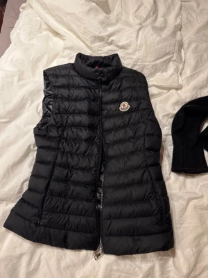 Svart dunväst från Moncler - Snygg svart dunväst från Moncler med quiltad design och hög krage. Västen har dragkedja framtill och Monclers ikoniska logotyp på bröstet. Perfekt för lager-på-lager och håller dig varm med lätt dunfyllning.