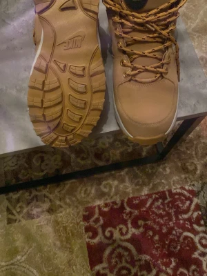 Vinter Nike skor gul och vit - Säljer ett par beige kängor från Nike med kraftig, mönstrad gummisula och guldfärgade öljetter. Skorna har snörning och svart vadderad kant upptill för extra komfort. Perfekta för dig som vill ha en streetig look och tåliga skor till hösten.