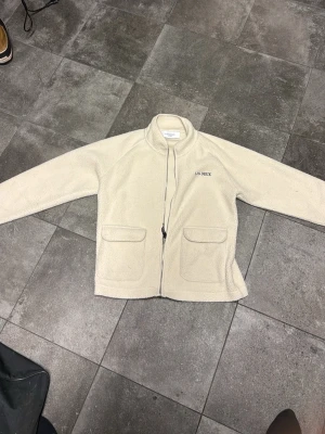 Beige teddyjacka från Les Deux - Mysig beige teddyjacka från Les Deux med dragkedja framtill och två stora fickor. Jackan har hög krage och broderad logga på bröstet. Perfekt för lager på lager och ger en avslappnad men stilren look.