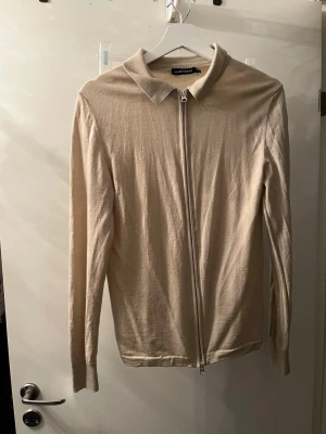 Beige ziptröja från J.Lindeberg - Stilren beige ziptröja från J.Lindeberg med klassisk krage och hel dragkedja framtill. Tillverkad i ett tunt, mjukt material som passar perfekt för lager på lager. Långärmad modell med enkel design som funkar till det mesta.