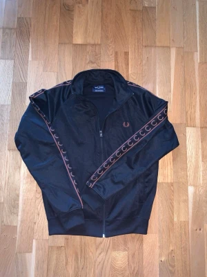 Svart track jacket från Fred Perry - Säljer en svart track jacket från Fred Perry med dragkedja framtill. Jackan har bruna detaljer och logga på bröstet samt brunt mönster längs ärmarna. Tillverkad i ett mjukt och stretchigt material, perfekt för en sportig och avslappnad stil. Tröjan har endast använts en gång. Pris kan diskuteras! Tveka inte på att höra av er vid eventuella frågor!