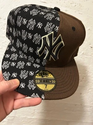Brun New Era 59FIFTY NY keps - Säljer en brun och svart New Era 59FIFTY keps med stort NY Yankees broderi framtill. Ena sidan är täckt av vita NY-loggor på svart botten, medan andra sidan är helbrun. Klassisk flat peak och broderade detaljer. Perfekt för dig som vill sticka ut med din stil.