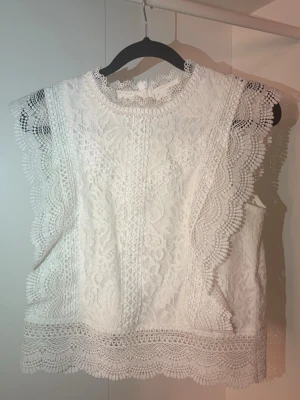 Vit spetsblus med scallopkant - Supersöt vit blus i spets med detaljerad scallopkant runt ärmhål och nederkant. Blusen är ärmlös och har rund halsringning samt knäppning bak i nacken. Perfekt för en chic och romantisk look. Materialet är genombruten spets i polyester.
