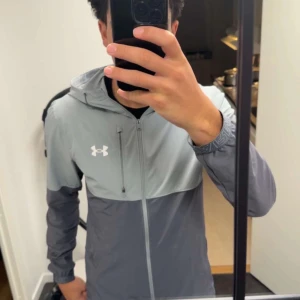 Grå vindjacka från Under Armour - Snygg  vindjacka från Under Armour i ljusgrått och mörkgrått. Jackan har huva, hel dragkedja framtill och elastiska muddar vid ärmsluten. Perfekt för blåsiga dagar och sportiga outfits. Under Armour-logga på bröstet ger en sportig vibe.
