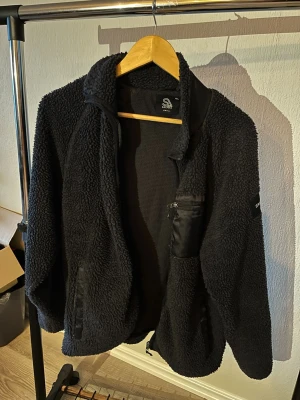 Svart teddyjacka från Saligia - Mysig svart teddyjacka från Saligia med dragkedja framtill, hög krage och detaljer i nylon vid fickor och bröst. Jackan har en avslappnad passform och är tillverkad i fluffigt teddy-material som håller dig varm under kyliga dagar.