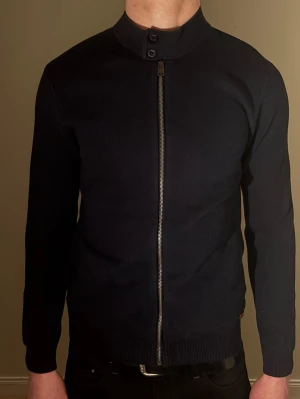 Mörkblå ziptröja med ståkrage - Snygg mörkblå tröja med hel dragkedja framtill och ståkrage med knappdetalj. Tröjan är i storlek m men är något tightare i passformen. Mycket bra skick!