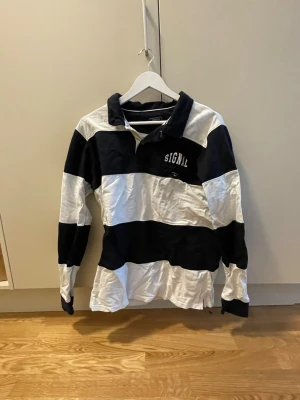 Randig rugbytröja  - Säljer en klassisk långärmad rugbytröja med breda svarta och vita ränder. Tröjan har krage, knappar vid halsen och broderad logga på bröstet. Tillverkad i mjuk bomull för skön känsla.