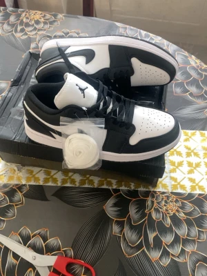 Svarta och vita Air Jordan 1 Low - Säljer ett par Air Jordan 1 Low i svart och vitt. Klassisk siluett med svart läder och vita detaljer, perforerad tåbox och ikonisk Jumpman-logga på plösen. Skorna kommer med extra vita skosnören. Perfekta för dig som gillar streetwear och sneakerskultur.