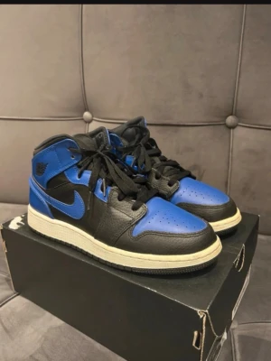 Air Jordan 1 Mid Svart/Blå - Snygga Air Jordan 1 Mid sneakers i svart och blått läder med vita sulor och svarta skosnören. Klassisk Nike Swoosh på sidan och Air Jordan-logga vid ankeln. Perfekta för dig som gillar streetwear och vill sticka ut med en ikonisk modell.