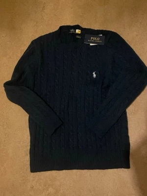  Polo Ralph Lauren - Kabelstickad mörkblå tröja från Polo Ralph Lauren med rund halsringning och den klassiska broderade logotypen i vitt på bröstet. Tröjan har långa ärmar och ribbade muddar. Perfekt för dig som gillar stilrena och tidlösa plagg.