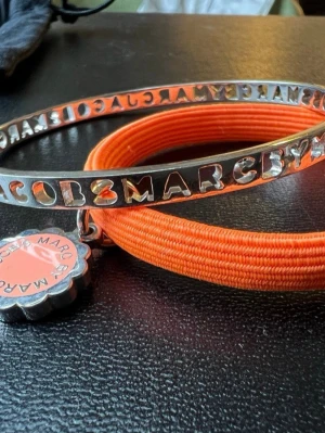 Marc Jacobs armbandsset – silver & korallorange - Snyggt armband från Marc by Marc Jacobs med orange elastiska band och ett silverfärgat metallband med utskuren logga. Armbandet har även en rund berlock i orange och silver med märkesnamnet. Perfekt accessoar för att addera färg till din outfit.
