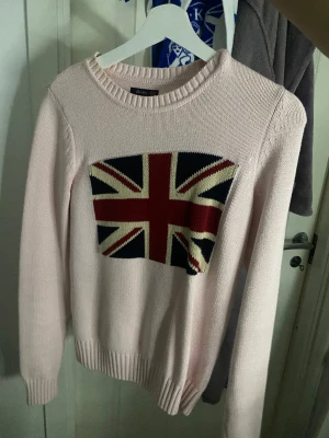 Ljusrosa stickad tröja med Storbritanniens flagga Crocker XS - Ljusrosa stickad tröja från Crocker med Union Jack-flagga i rött, blått och vitt på bröstet. Tröjan har rund halsringning, ribbade muddar och är i ett mjukt stickat material. Perfekt för dig som gillar statement-plagg och snygga detaljer. Liknande Ralph Lauren