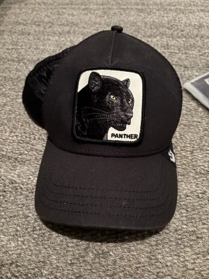 Svart keps med panterpatch - Snygg svart keps från Goorin Bros med mesh baktill och en broderad patch framtill med en svart panter och texten 'PANTHER'. Justerbar snapback och böjd skärm. Perfekt för dig som gillar djurmotiv och streetwear.