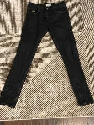 Svarta skinny jeans från Lacoste - Säljer ett par svarta skinny jeans från Lacoste med klassisk femficksmodell och det ikoniska krokodilmärket på fickan. Jeansen har knappgylf och smal passform som sitter tight hela vägen ner. Perfekta till en streetwear-look.