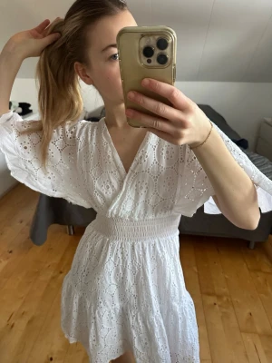 Vit kort klänning med broderade detaljer - Superfin vit kort klänning med broderade hålmönster och omlott-v-ringning. Klänningen har vida ärmar och smock i midjan för en bekväm passform. Perfekt för varma dagar och framförallt STUDENT🥹