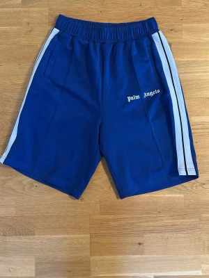 Blå shorts från Palm Angels - Snygga blå shorts från Palm Angels med vita sidoränder och logga på vänster ben. Shortsen har elastisk midja och två sidofickor. Perfekta för en sportig och avslappnad stil.
