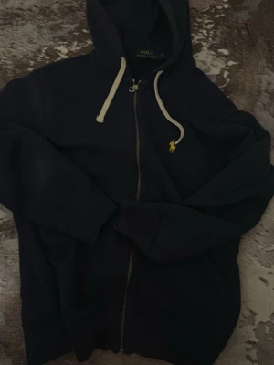 Marinblå hoodie från Polo Ralph Lauren - Snygg marinblå hoodie från Polo Ralph Lauren med dragkedja framtill och vit dragsko i huvan. Klassisk gul broderad logga på bröstet. Tillverkad i mjukt bomullsmaterial som känns skönt mot huden. Perfekt för en avslappnad och stilren look.