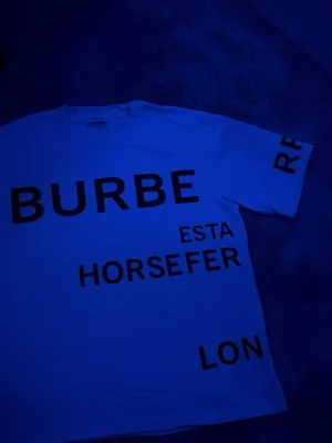 Vit t-shirt med svart text Burberry - Säljer en vit t-shirt från Burberry med stora svarta bokstäver och texttryck både fram och bak. T-shirten har klassisk passform och korta ärmar. Perfekt för dig som gillar statement-plagg och vill sticka ut med en ikonisk design.