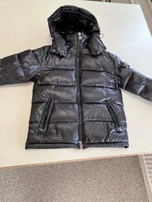 Svart dunjacka från Moncler med huva - Snygg svart dunjacka från Moncler med avtagbar huva och två dragkedjeförsedda fickor framtill. Jackan har klassisk puffer-design och Moncler-logga på ärmen. Perfekt för kalla vinterdagar och tillverkad i glansigt material som ger en exklusiv look.