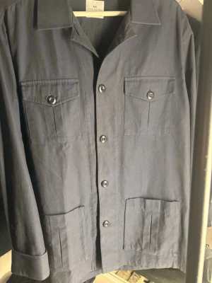 Mörkblå overshirt från John Henric - Stilren mörkblå overshirt från John Henric med fyra fickor framtill, två med lock och knapp samt två utan lock. Klassisk krage och knäppning med svarta knappar. Tillverkad i ett kraftigare bomullstyg som ger en snygg och avslappnad look. Aldrig använd 