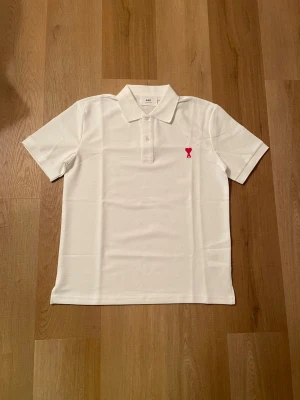 Vit piké från Ami Paris med rött hjärta - Klassisk vit pikétröja från Ami Paris med rött broderat hjärta och bokstaven A på bröstet. Skjortan har korta ärmar, krage och knappar framtill. Tillverkad i mjuk bomull för en clean och stilren look.