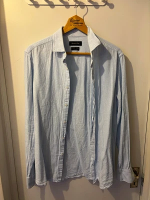 Ljusblå skjorta från Morris - Klassisk ljusblå skjorta från Massimo Dutti med diskret rutigt mönster. Skjortan har lång ärm, knappar framtill och är tillverkad i mjuk bomull. Perfekt för en clean och stilren look.