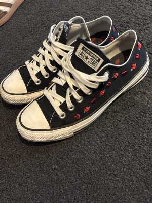 Converse All Star svarta - Säljer ett par svarta Converse All Star sneakers med vita sulor och snören. Skorna har broderade röda rosor på sidorna som ger en unik och personlig touch. Klassisk låg modell i canvas med gummitå och logga på plösen.