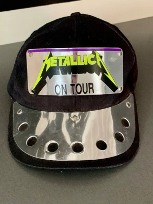 Y2K • Vintage • Metallica - ON TOUR Black Era • Snapback - Original vintage Metallica “ON TOUR” snapback-keps från sent 80-tal / tidigt 90-tal. Klassisk Metallica-relaterad vintage-keps från bandets storhetstid. Kepsen har den ikoniska neon-gröna Metallica-loggan monterad på en nitad metallplatta och formgjuten skärm – en design som är typisk för slutet av 80-talet och början av 90-talet och väldigt ovanlig.  En original vintage-produkt från perioden, inte någon nytillverkad replika eller modern kopia.